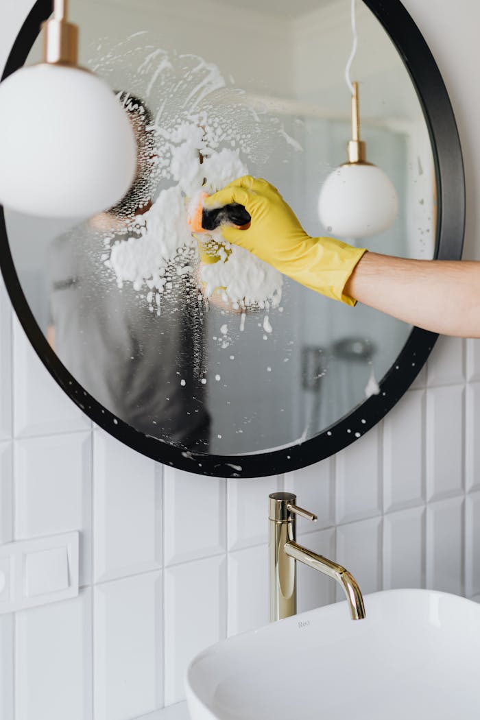 about-01 Anonymous housekeeper in yellow rubber gloves wiping round mirror by sponge in bathroom